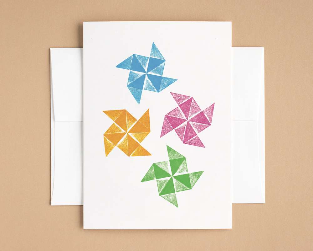 Four origami pinwheels – colored blue, orange, magenta, and green – are scattered across the surface of a white vertical card. The card lies on top of a white envelope, which lies on a brown backdrop.